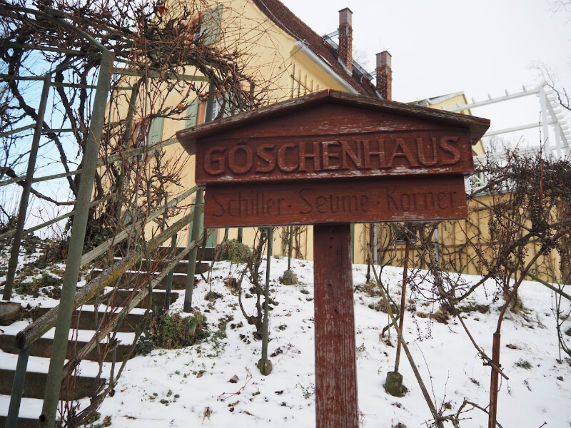 Exterior of the Göschen House museum in Grimma, a two-story building with a red and white facade, large windows, and a sign above the entrance; the scene is set on a sunny day with a clear blue sky